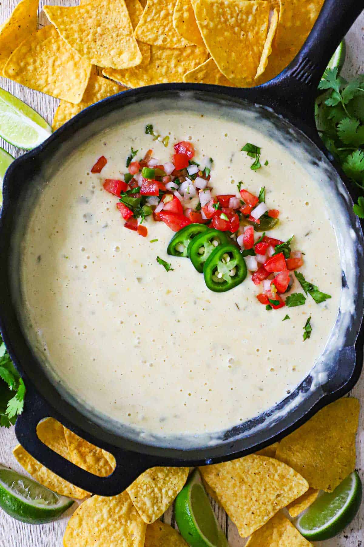 An overhead view of a cast-iron skillet filled with restaurant-style white queso (blanco) that has been garnished with pico de gallo, sliced jalape&ntilde;os, and chopped cilantro.