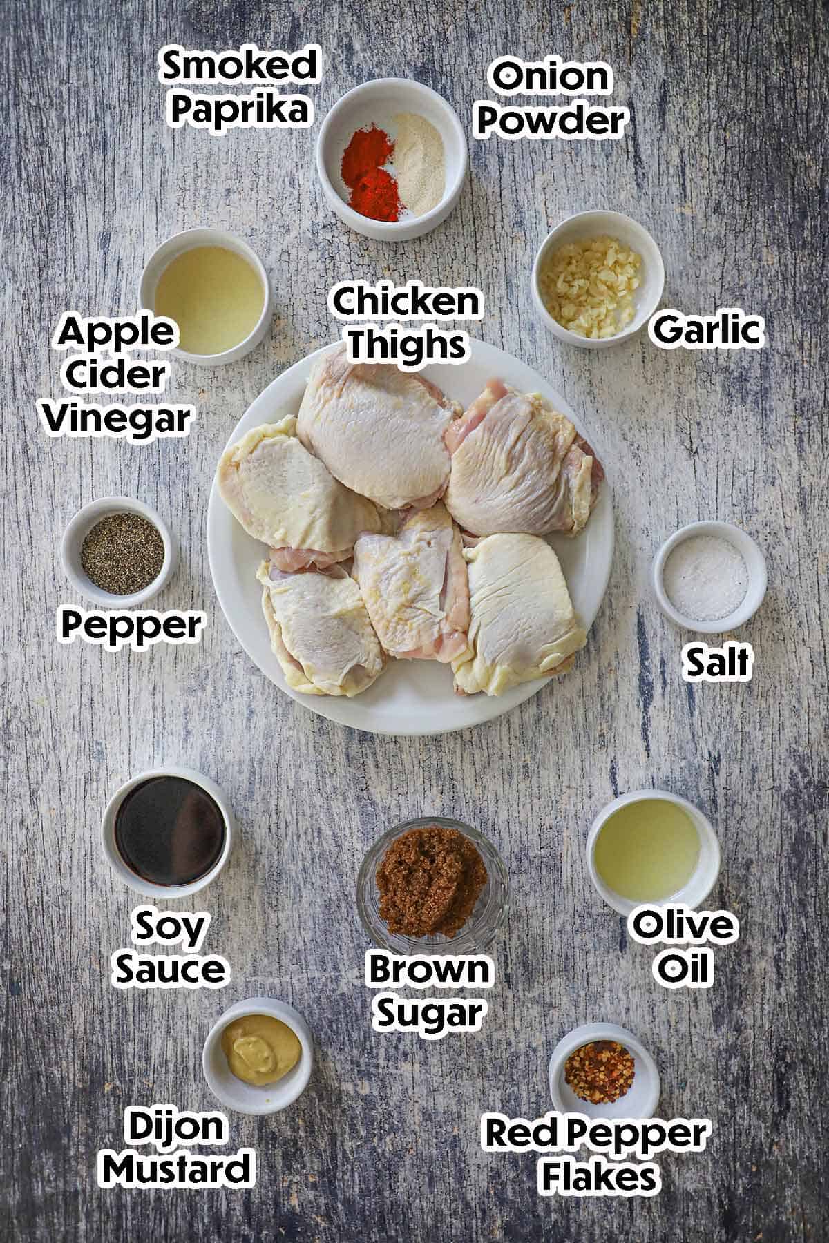 An arrangement of ingredients for baked brown sugar garlic chicken on a grey wooden cutting board including raw chicken thighs, vinegar, brown sugar, soy sauce, Dijon mustard, oil, garlic, and seasonings.