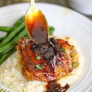 A person using a spoon to drizzle sauce over the top of a baked brown sugar garlic chicken thigh that is resting on a bed of mashed potatoes.