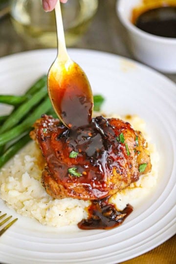A person using a spoon to drizzle sauce over the top of a baked brown sugar garlic chicken thigh that is resting on a bed of mashed potatoes.