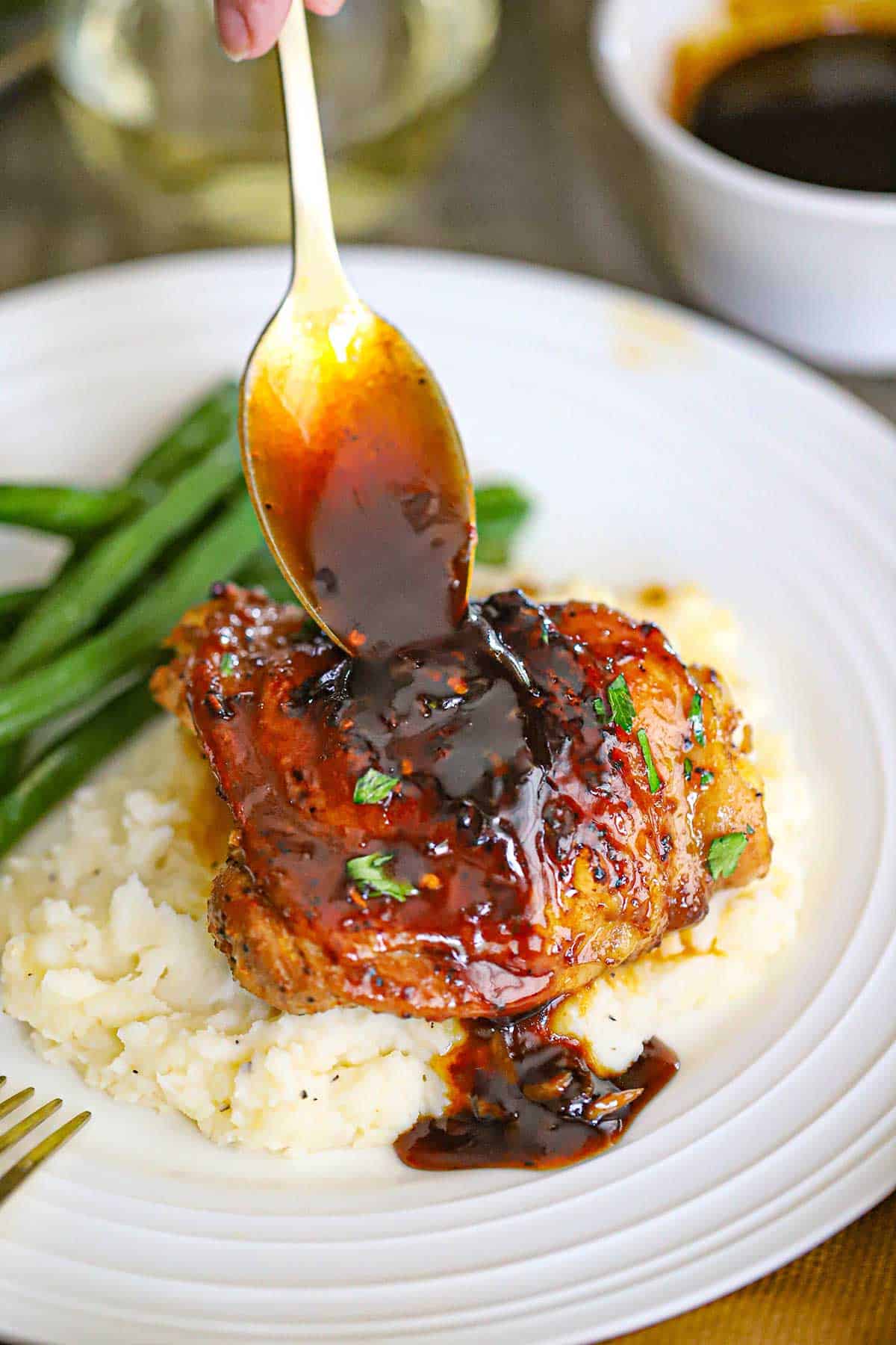 A person using a spoon to drizzle sauce over the top of a baked brown sugar garlic chicken thigh that is resting on a bed of mashed potatoes.