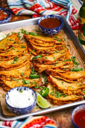 A large silver sheet pan filled with two rows of overlapping crispy baked chicken tacos garnished with chopped cilantro.