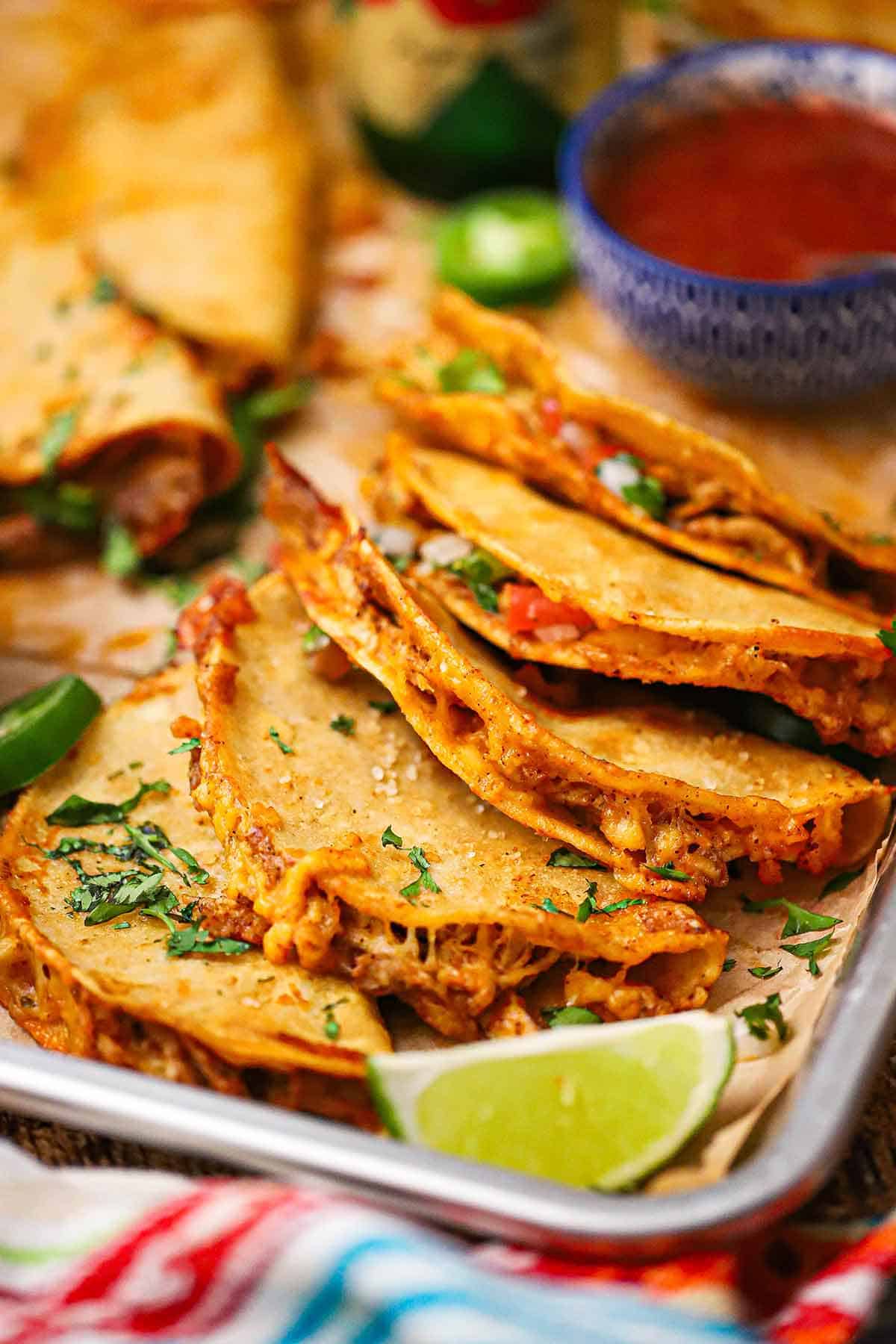 A close-up view of a silver trey filled with Crispy Baked Chicken Tacos stacked against one another with a lime wedge and a bowl of enchilada sauce nearby.