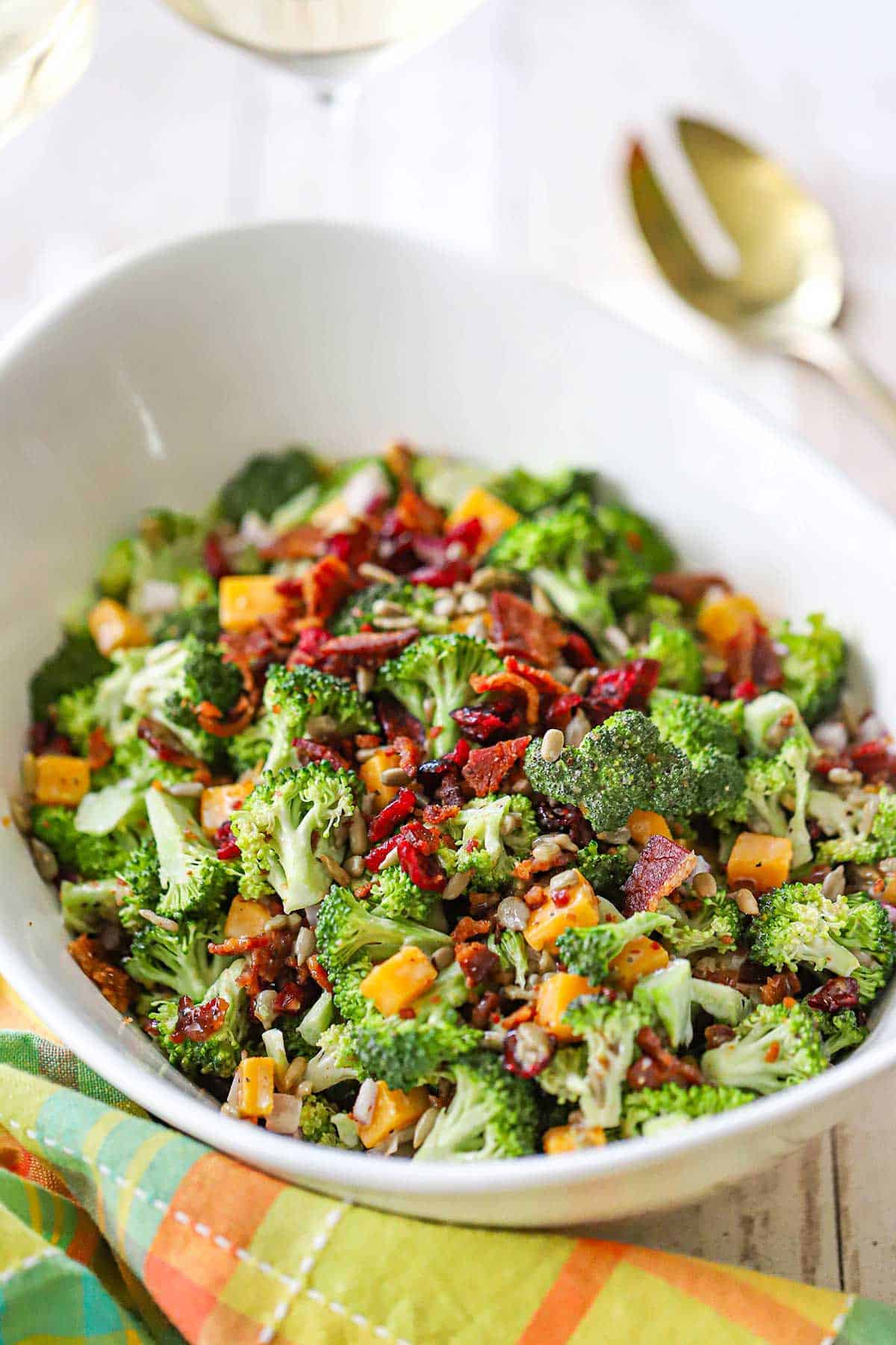 A close-up view of a white serving bowl filled with broccoli salad with bacon and topped with dried cranberries, cubed cheddar cheese, and sunflower seeds.