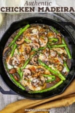 An overhead view of a large cast-iron skillet filled with chicken madeira topped with sautéed mushrooms and asparagus.
