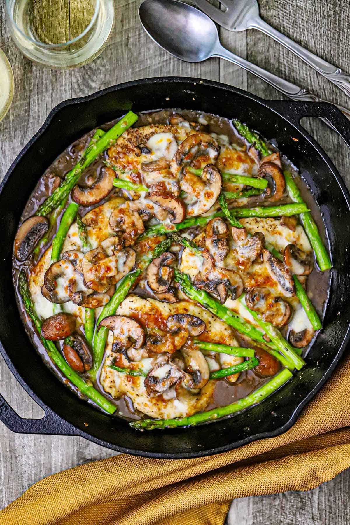 An overhead view of a large cast-iron skillet filled with chicken madeira topped with sautéed mushrooms and asparagus.