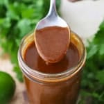 A person using a spoon to hold up a spoonful of easy homemade red enchilada sauce from a jar filled with the same.