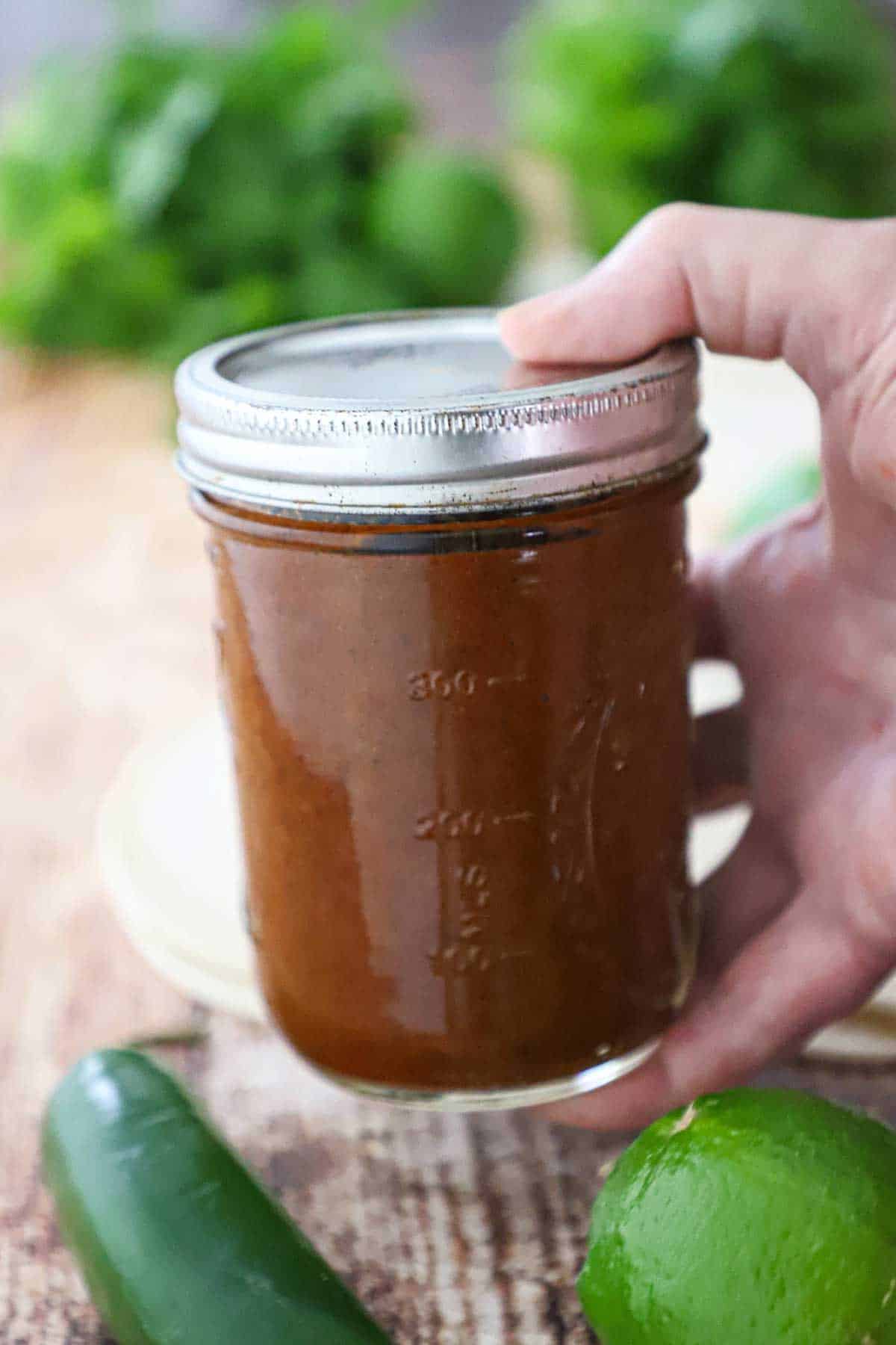 A person holding a Mason jar filled with Easy Homemade Red Enchilada Sauce between his thumb and fingers.