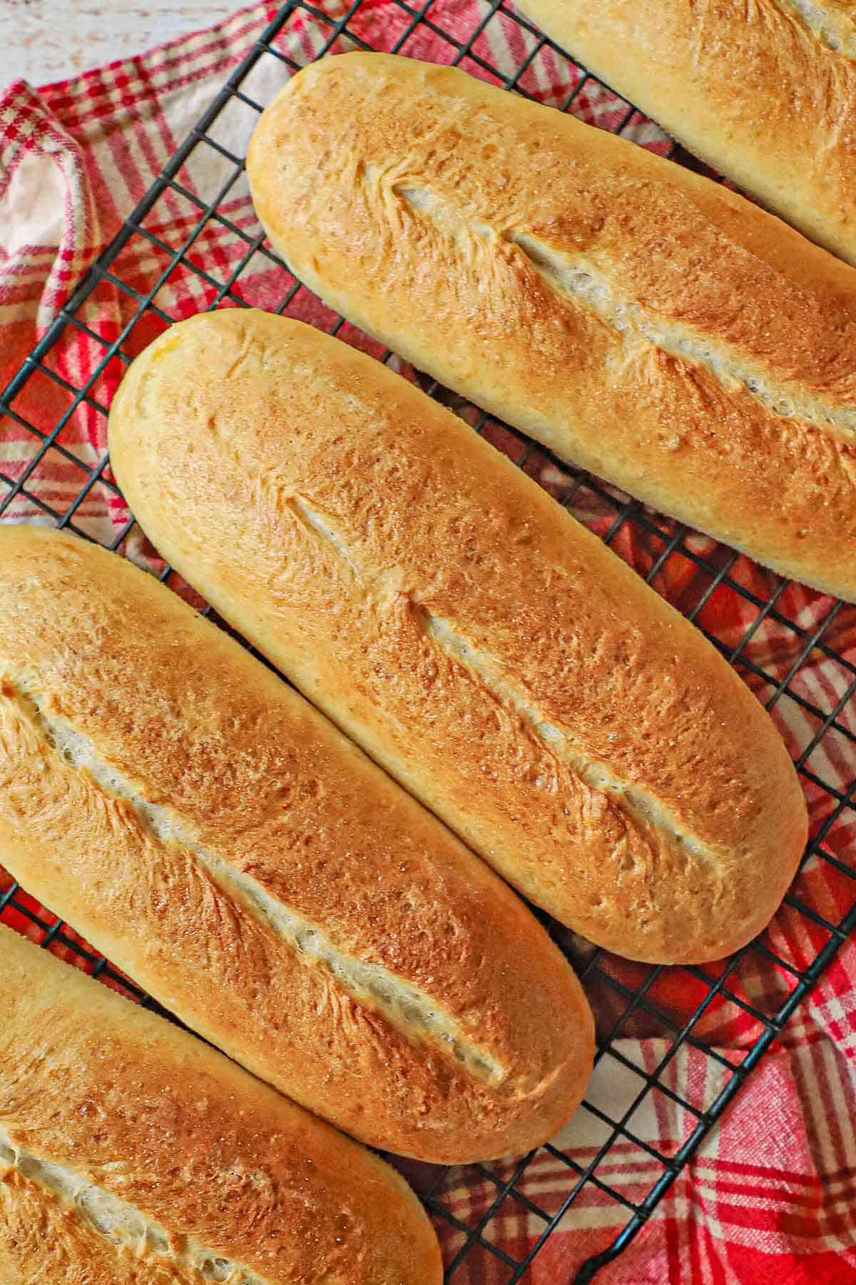 An overhead view of a row of homemade Italian rolls resting on a baking rack over a red and white linen napkin.