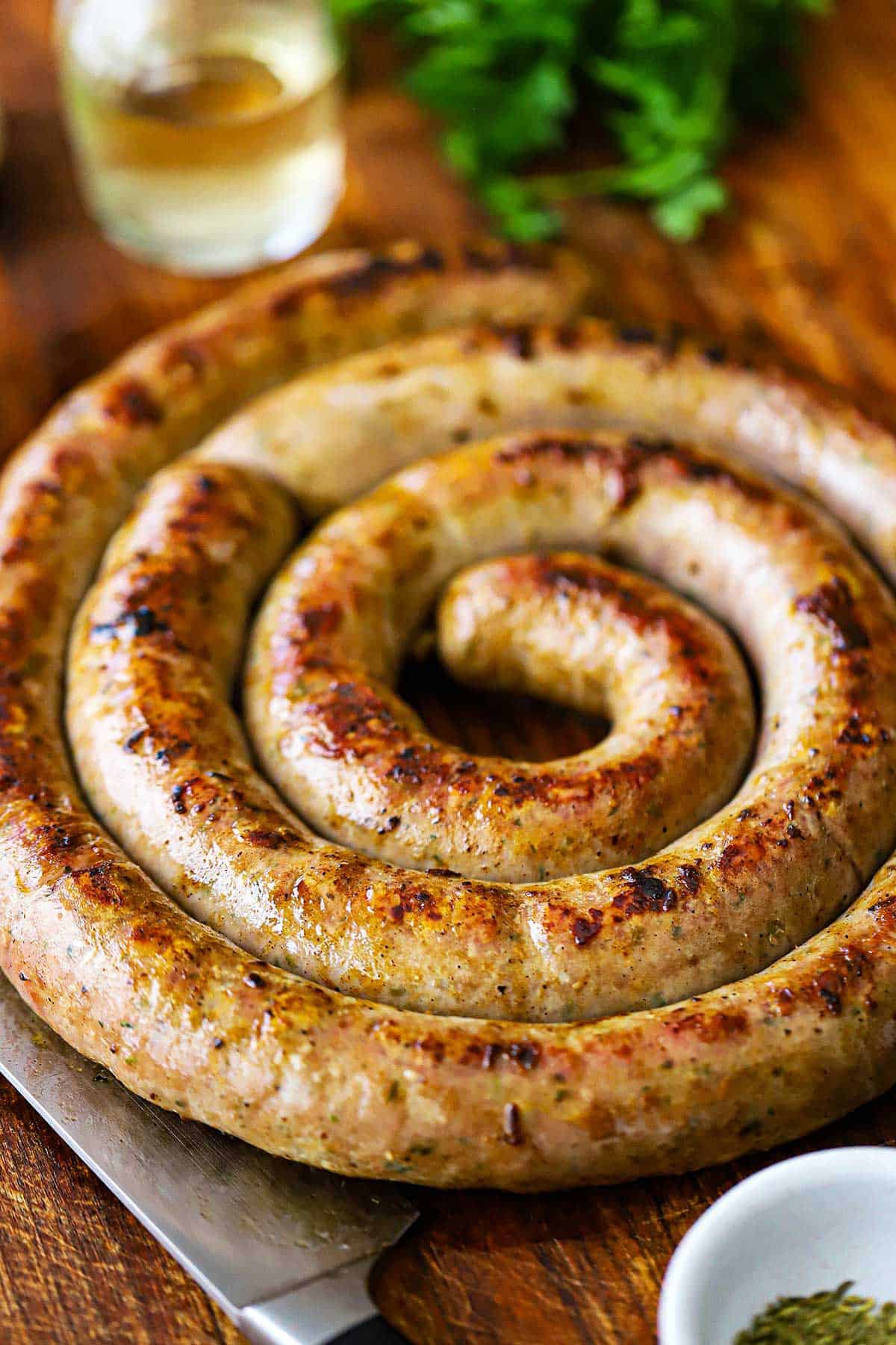 A straight-on view of a circular rope of homemade Italian sausage that has been seared and cooked and is resting on a wooden cutting board with small bowls of seasoning and Italian parsley nearby.