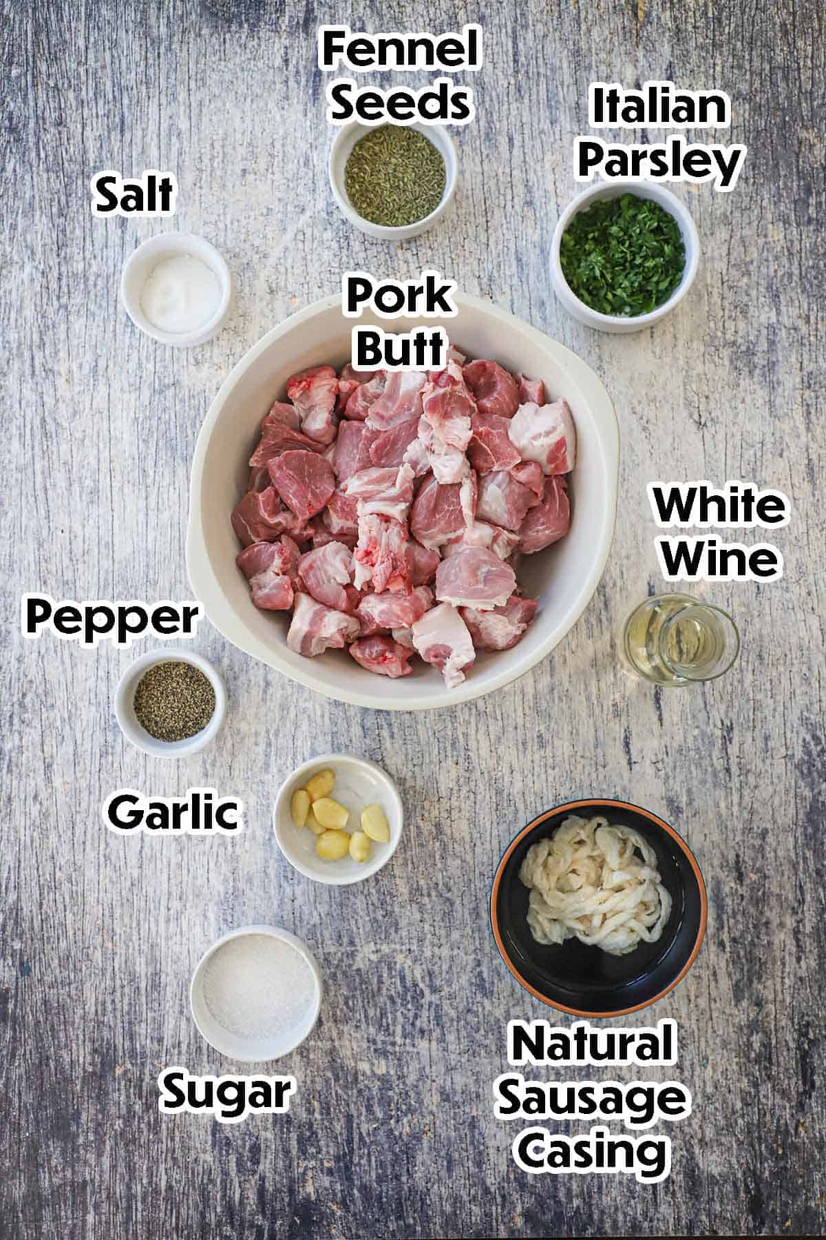 An arrangement of ingredients for Homemade Italian Sausage on a grey wooden background including cubed pork butt, natural sausage casings, garlic, sugar, and seasonings.
