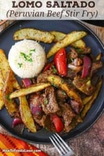 An overhead view of a serving of Classic Lomo Saltado, Peruvian Steak and Potatoes Stir-Fry, on a black dinner plate with a mound of Peruvian Garlic Rice on the plate as well.