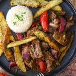 An overhead view of a serving of Classic Lomo Saltado, Peruvian Steak and Potatoes Stir-Fry, on a black dinner plate with a mound of Peruvian Garlic Rice on the plate as well.