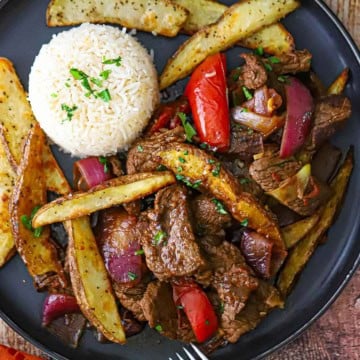 An overhead view of a serving of Classic Lomo Saltado, Peruvian Steak and Potatoes Stir-Fry, on a black dinner plate with a mound of Peruvian Garlic Rice on the plate as well.