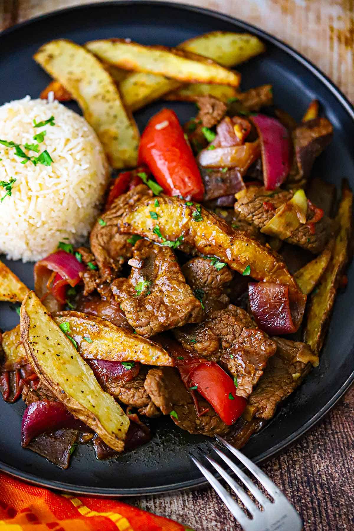 An overhead view of a serving of Classic Lomo Saltado, Peruvian Steak and Potatoes Stir-Fry, on a black dinner plate with a mound of Peruvian Garlic Rice on the plate as well.