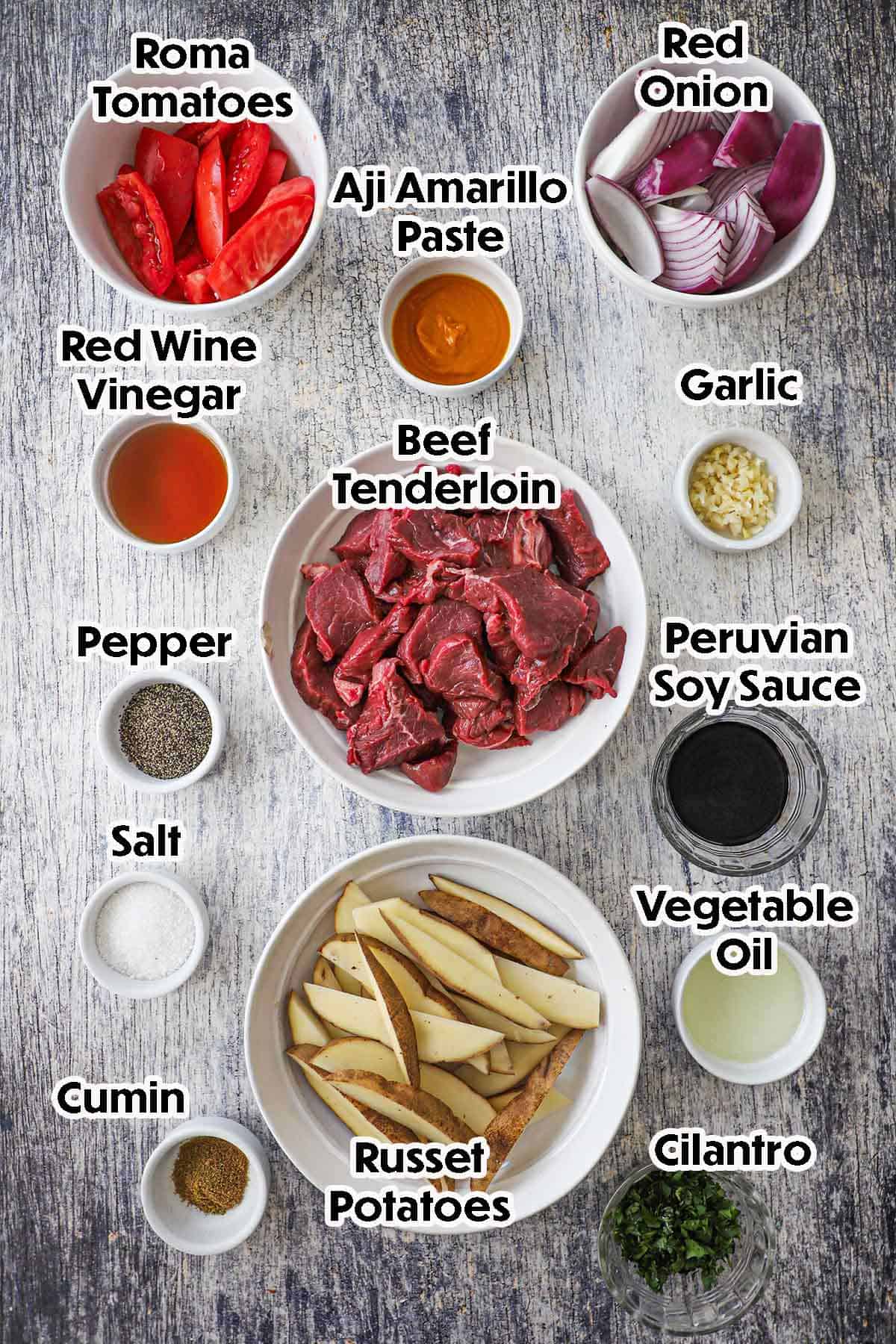 An arrangement of ingredients for Classic Lomo Saltado on a grey wooden background including beef tenderloin, pepper paste, roma tomatoes, red onion, potato wedges, garlic, oil, and seasonings.