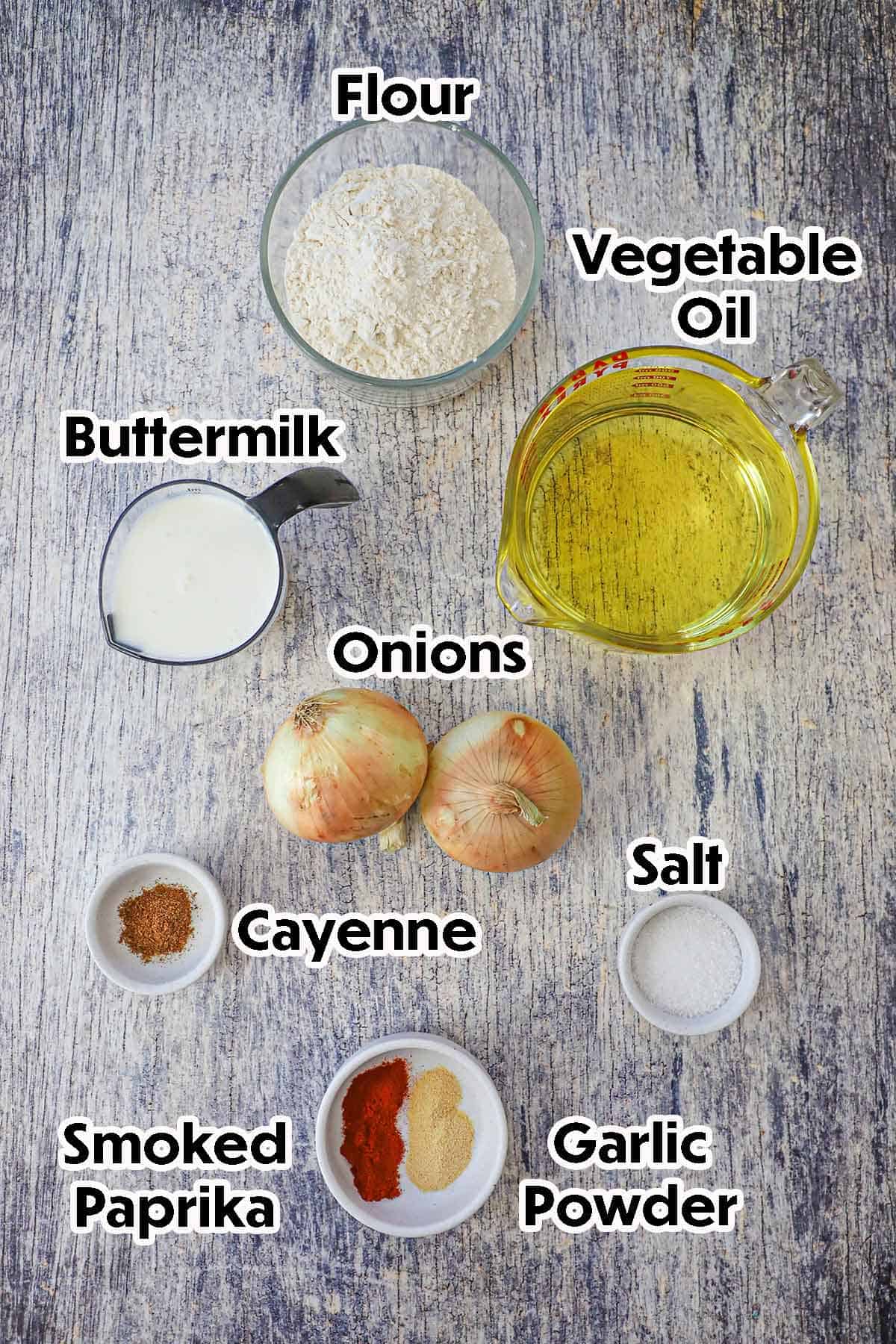 An arrangement of ingredients for steakhouse onion rings on a grey wooden background including sliced onions, buttermilk, flour, oil, and seasonings. 