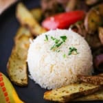 A close-up view of Peruvian Garlic Rice that has been molded into a small dome and is resting on a black dinner plate next to a serving of Peruvian Beef and Potato Stir-Fry.