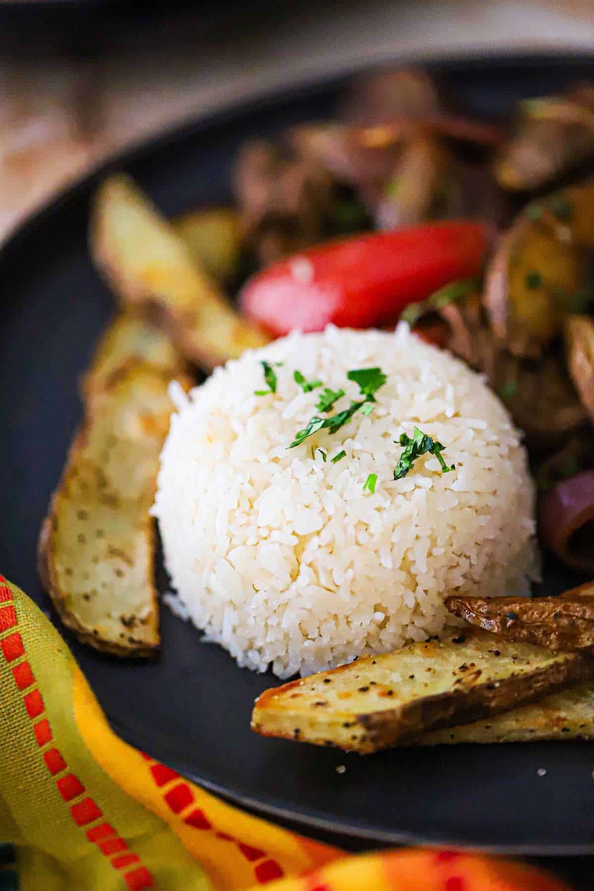 A close-up view of Peruvian Garlic Rice that has been molded into a small dome and is resting on a black dinner plate next to a serving of Peruvian Beef and Potato Stir-Fry.