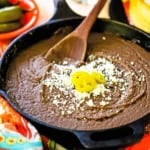 A close-up view of a large black cast-iron skillet filled with authentic homemade refried beans topped with crumbled Cotija cheese and three slices of pickled jalapeños.