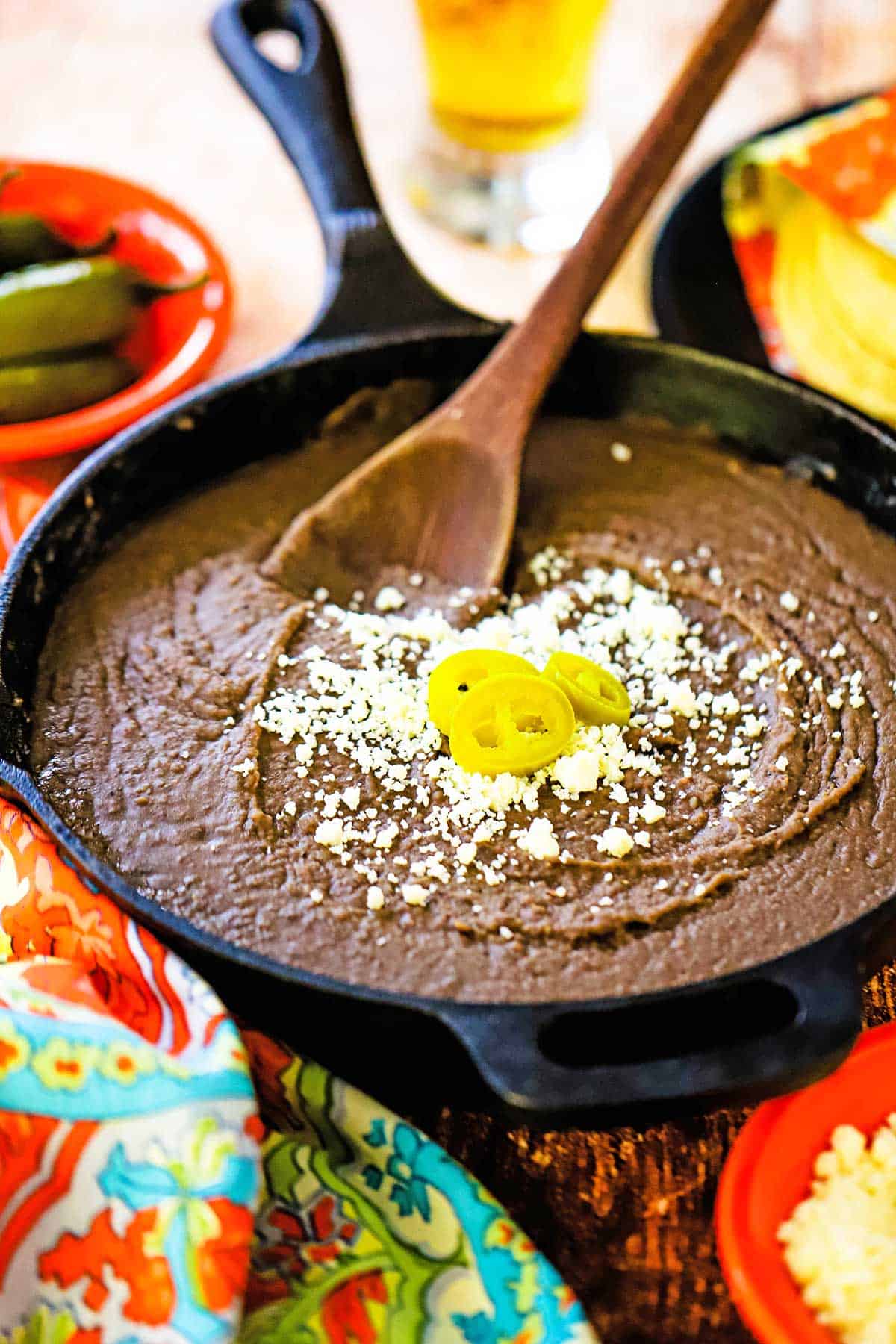 A large black cast-iron skillet filled with authentic homemade refried beans topped with crumbled Cotija cheese and three slices of pickled jalape&ntilde;os.