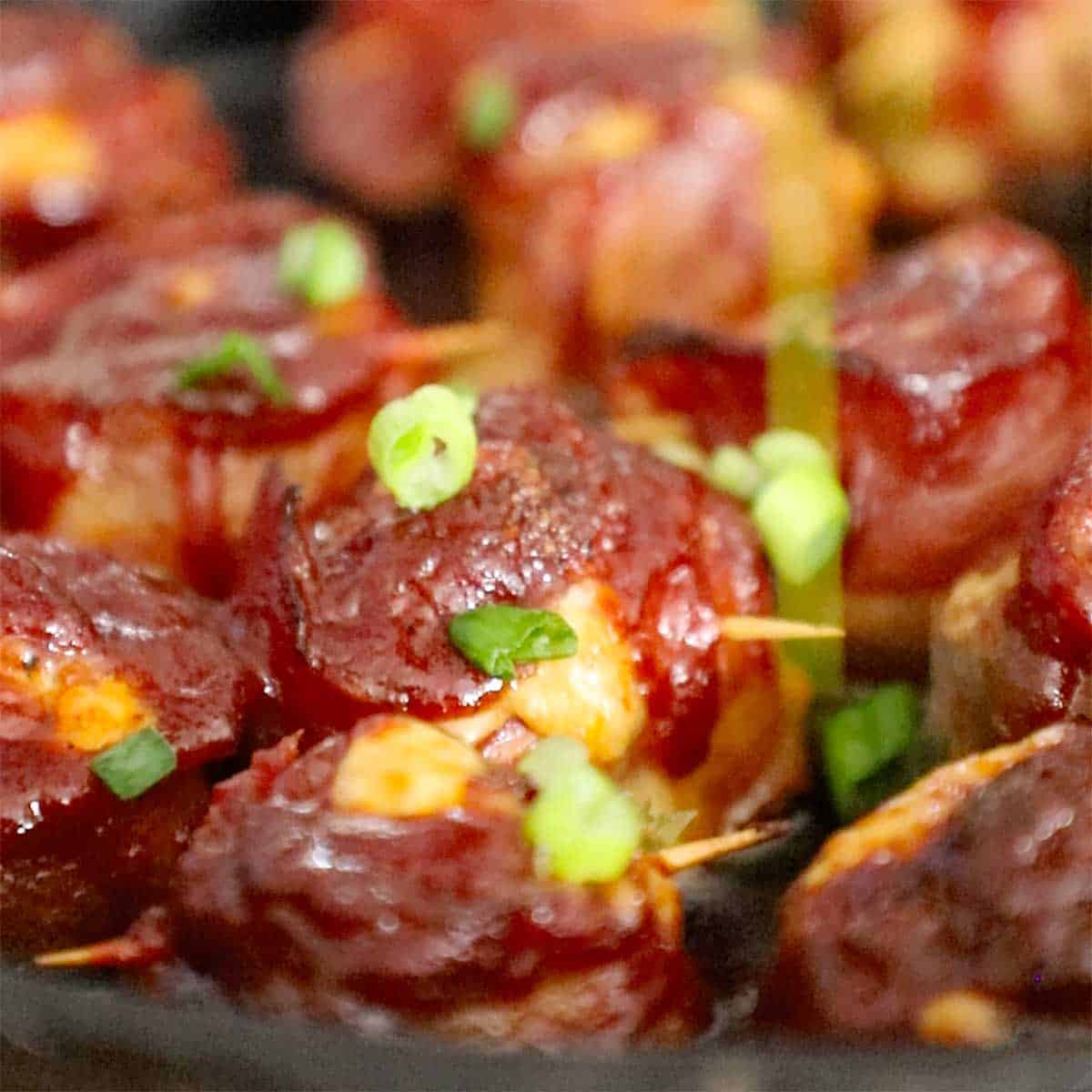 A person sprinkling chopped scallions over the tops of smoked bacon-wrapped meatballs topped with BBQ sauce.