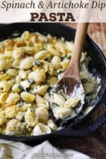 A wooden spoon resting in a large cast-iron skillet filled with Spinach Artichoke Dip Pasta.