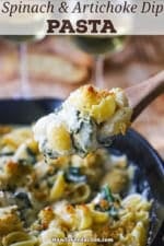 A person using a large wooden spoon to lift a serving of Spinach Artichoke Dip Pasta from a skillet filled with the same.