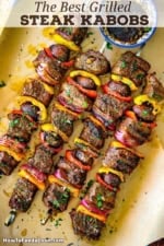 A close-up overhead view of four metal skewer filled with grilled steak kabobs of chunks of beef tenderloin, vegetables, and mushrooms topped with a garlic and balsamic vinaigrette.