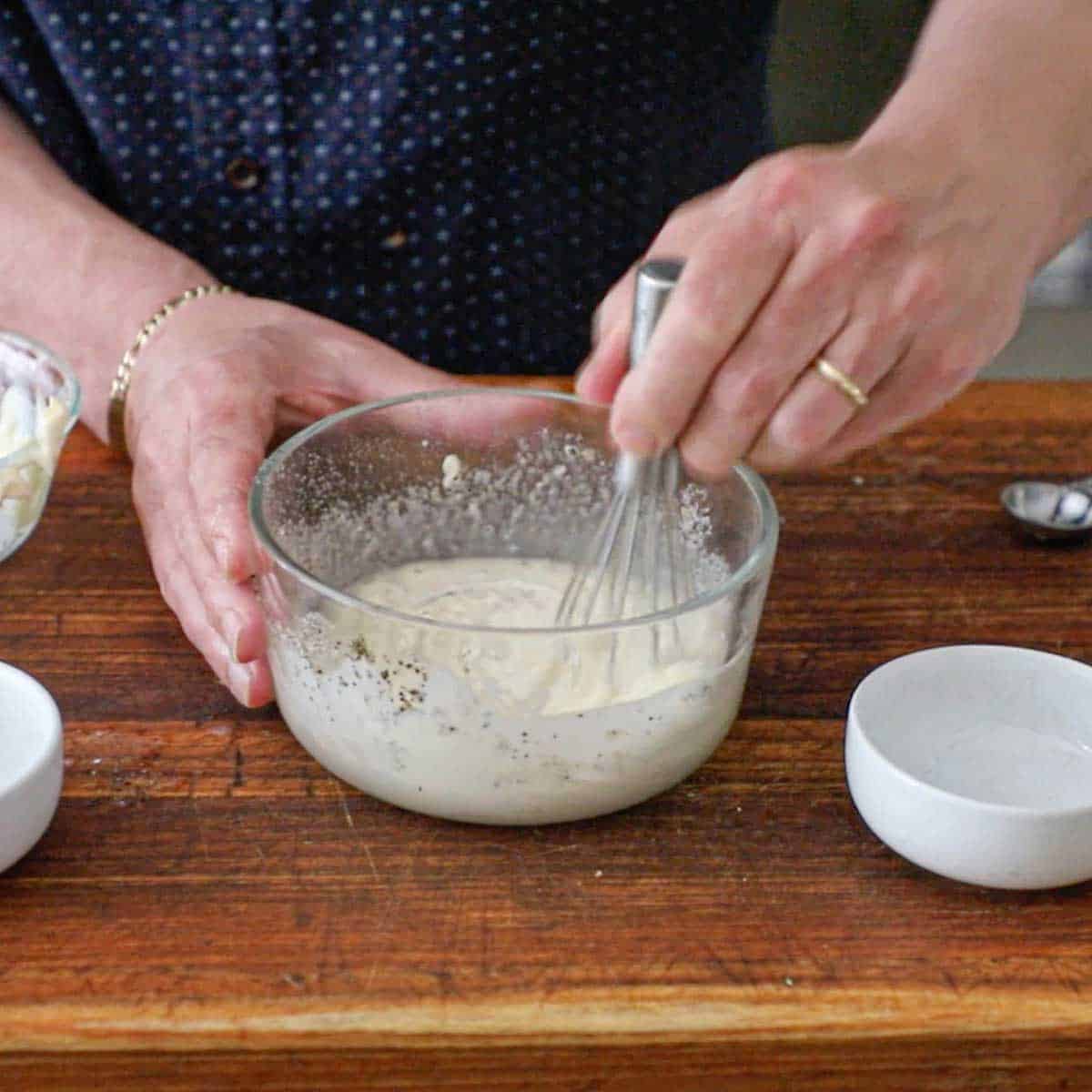 A person using a small whisk to combine a mayonnaise and vinegar dressing in glass bowl resting on a wooden cutting board.