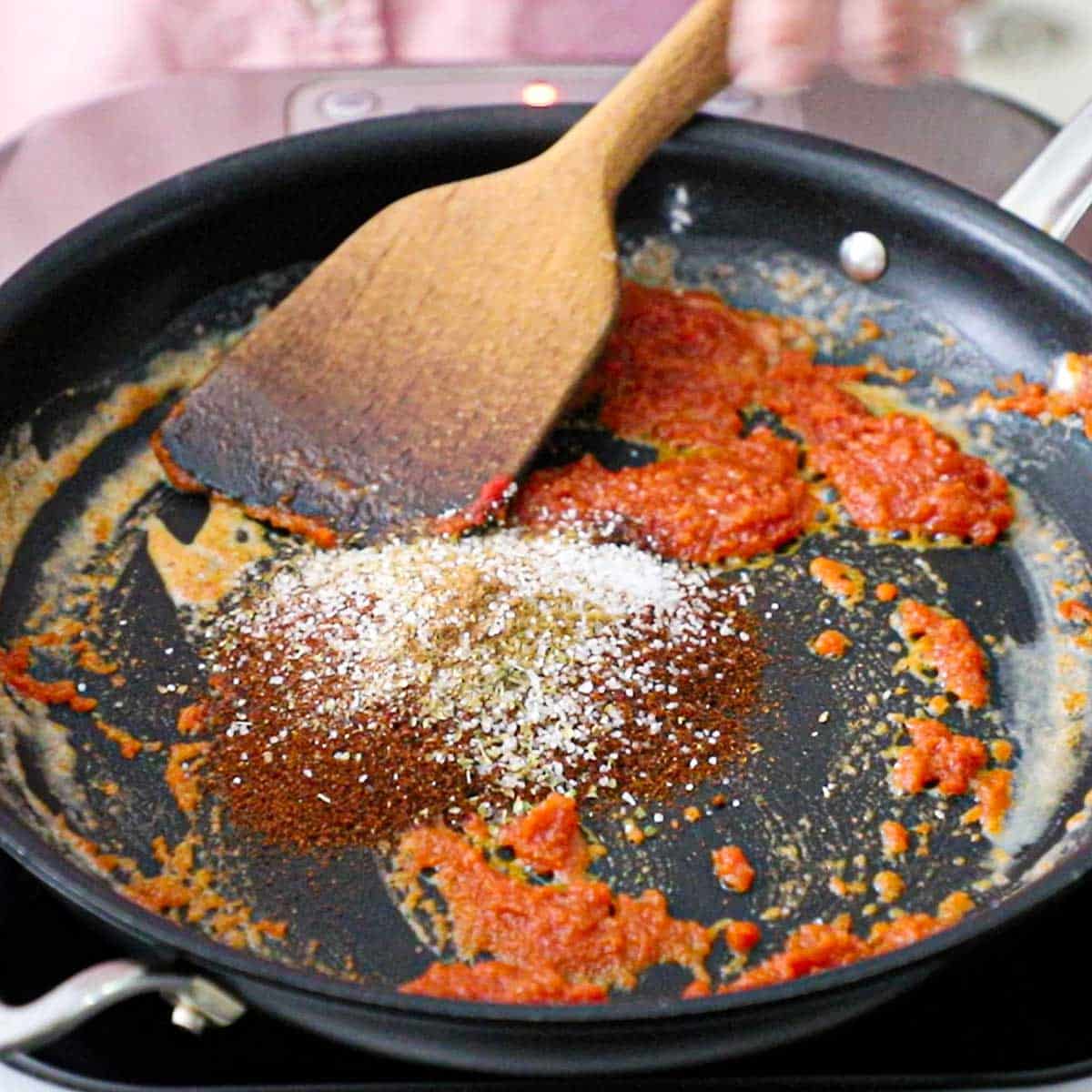 A person using a wooden spatula to stir chili powder and other seasonings into a skillet that contains tomato paste that has been cooked with a roux.