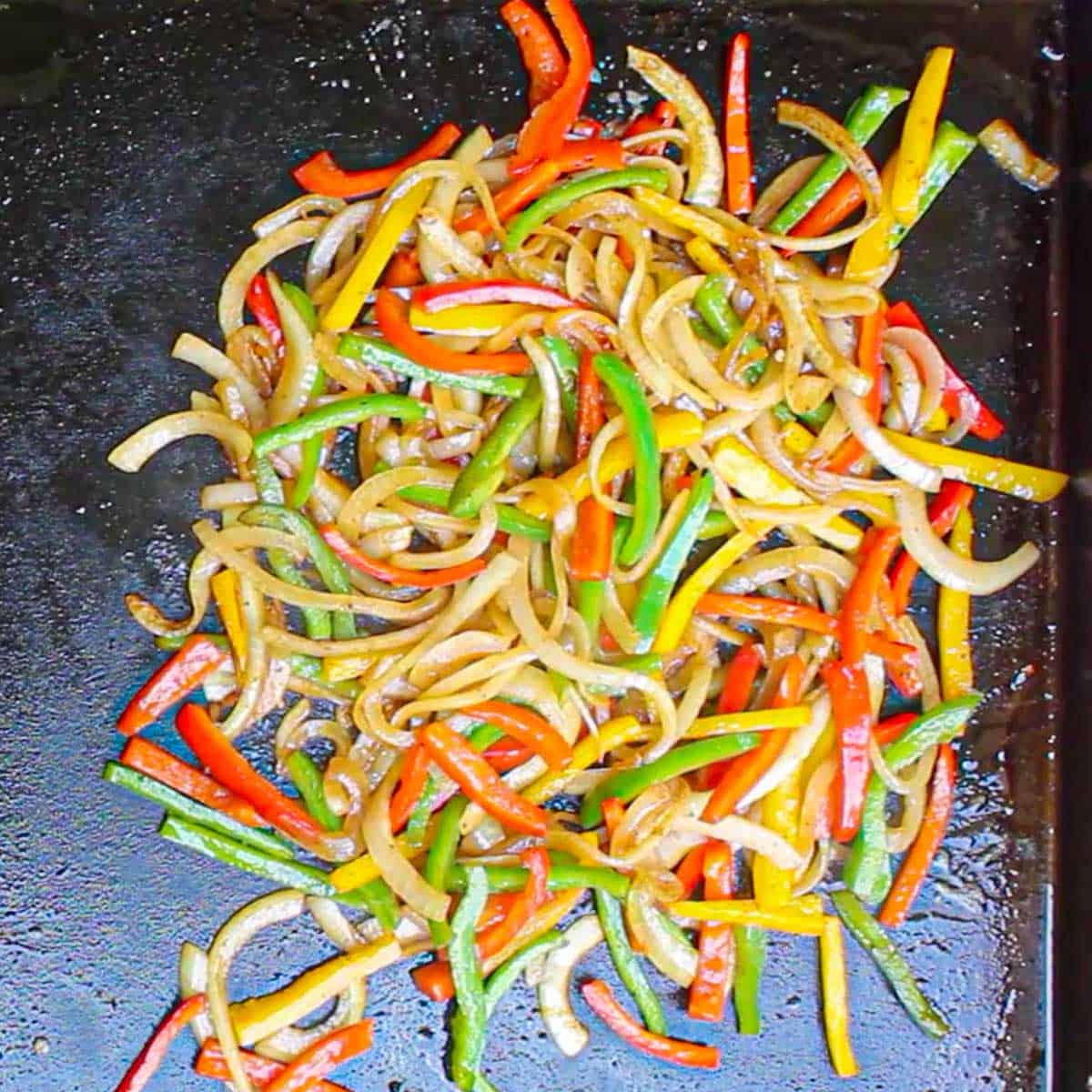 An overhead view of strips of onion, and colorful strips of bell peppers being sautéed in oil on a large outdoor griddle.
