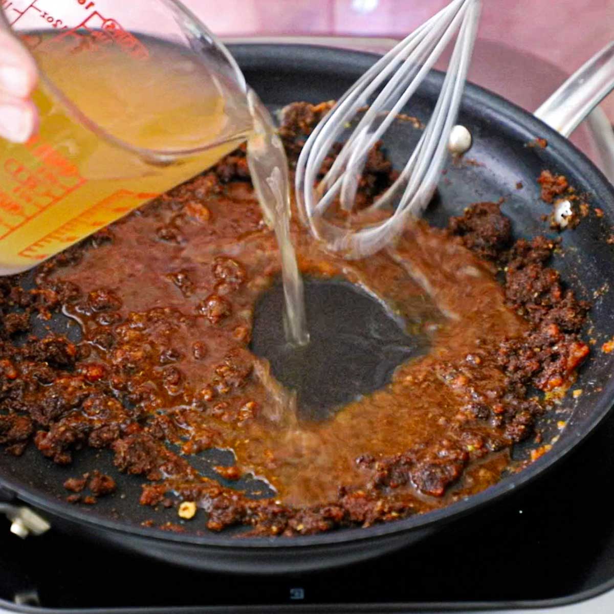 A person using a whisk to stir chicken broth that is being poured into an enchilada sauce roux that is simmering in a large black skillet.