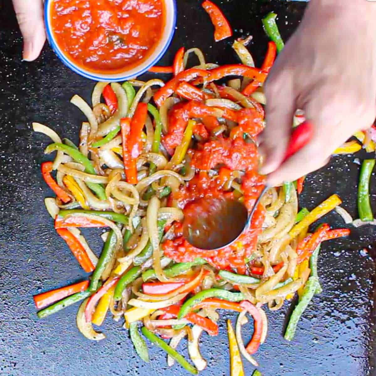 A person using a small ladle to drizzle marinara sauce of the top of sautéed peppers and onions simmering on a large outdoor griddle.