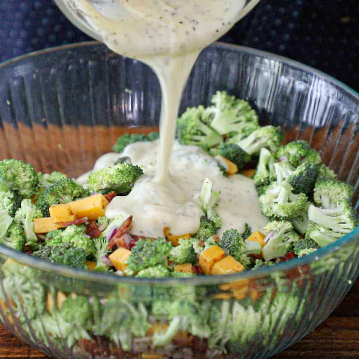 A person pouring a creamy dressing over the top of the components of broccoli salad with bacon in a large glass bowl.