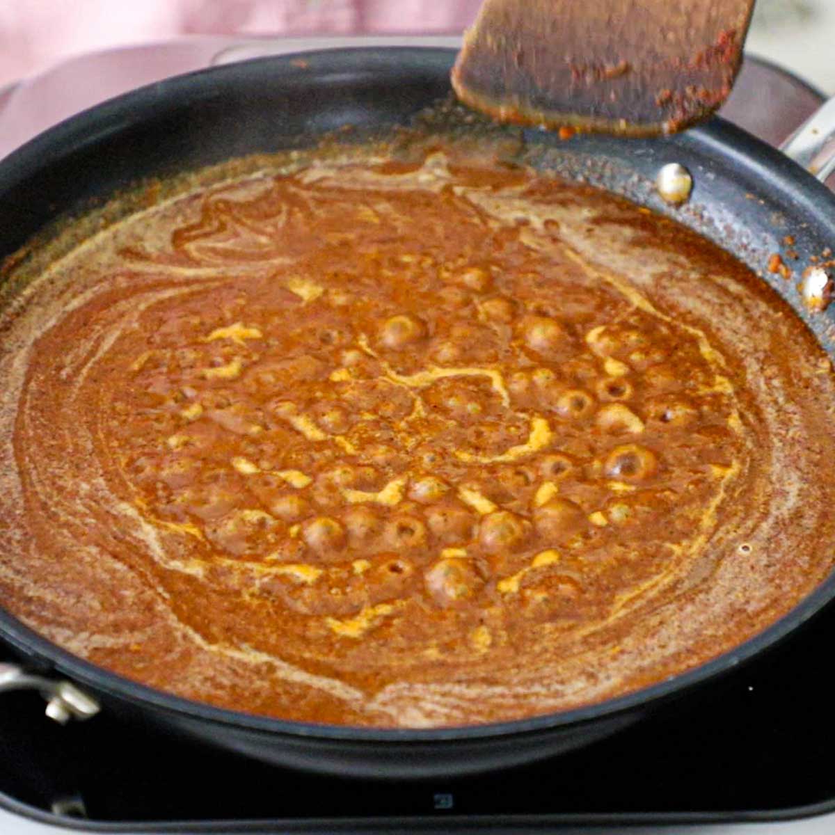 A person using a wooden spatula to stir gurgling red enchilada sauce in a large black non-stick skillet.