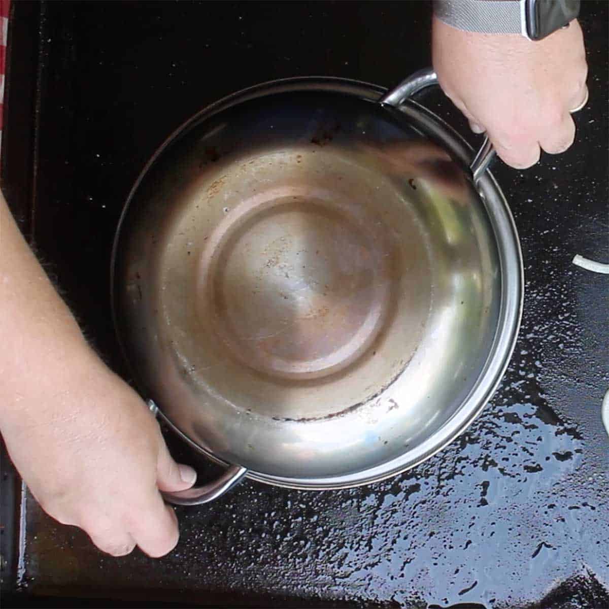 A person placing a large silver pot over a cooked rope of Italian sausage on a large outdoor griddle.