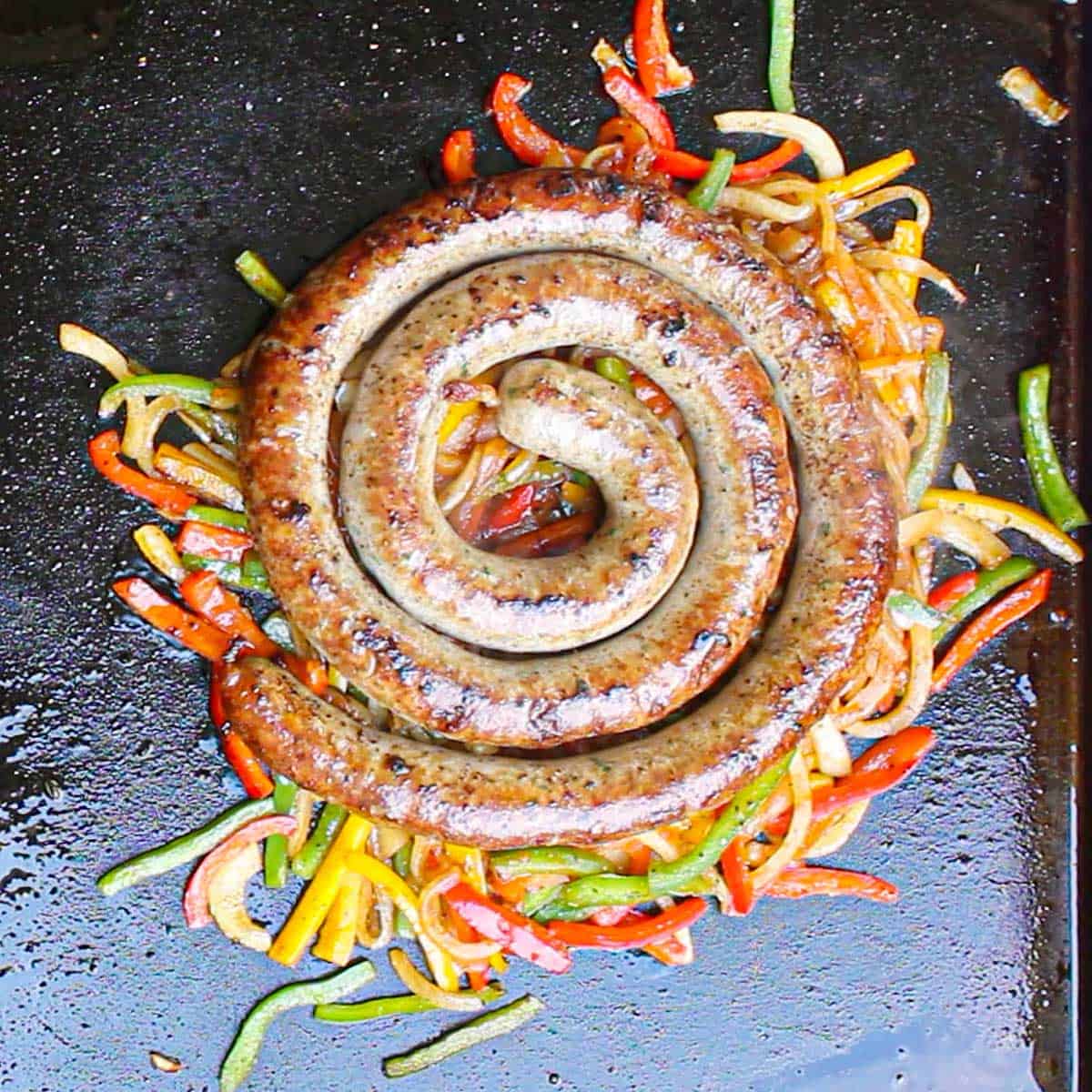 An overhead view of a fully cooked coiled rope of Italian sausage resting on a bed of sautéed strips of onions and bell peppers on an outdoor griddle.