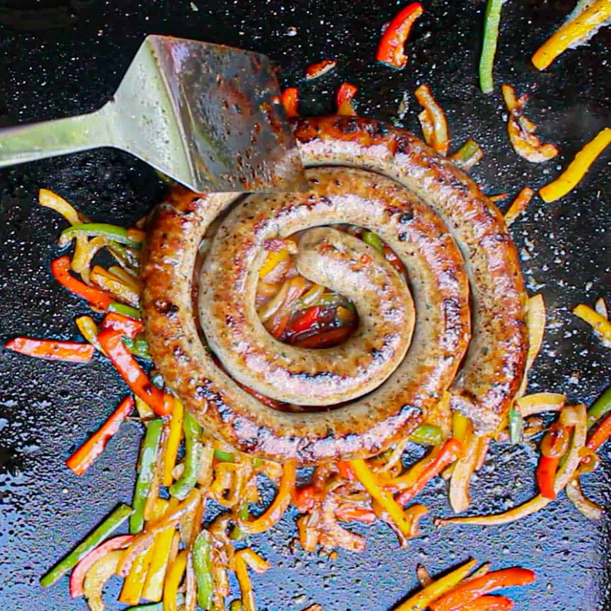A person using a large metal spatula to cut a large link from a fully cooked rope of Italian sausage that is simmering on a bed of sautéed peppers and onions on an outdoor griddle.