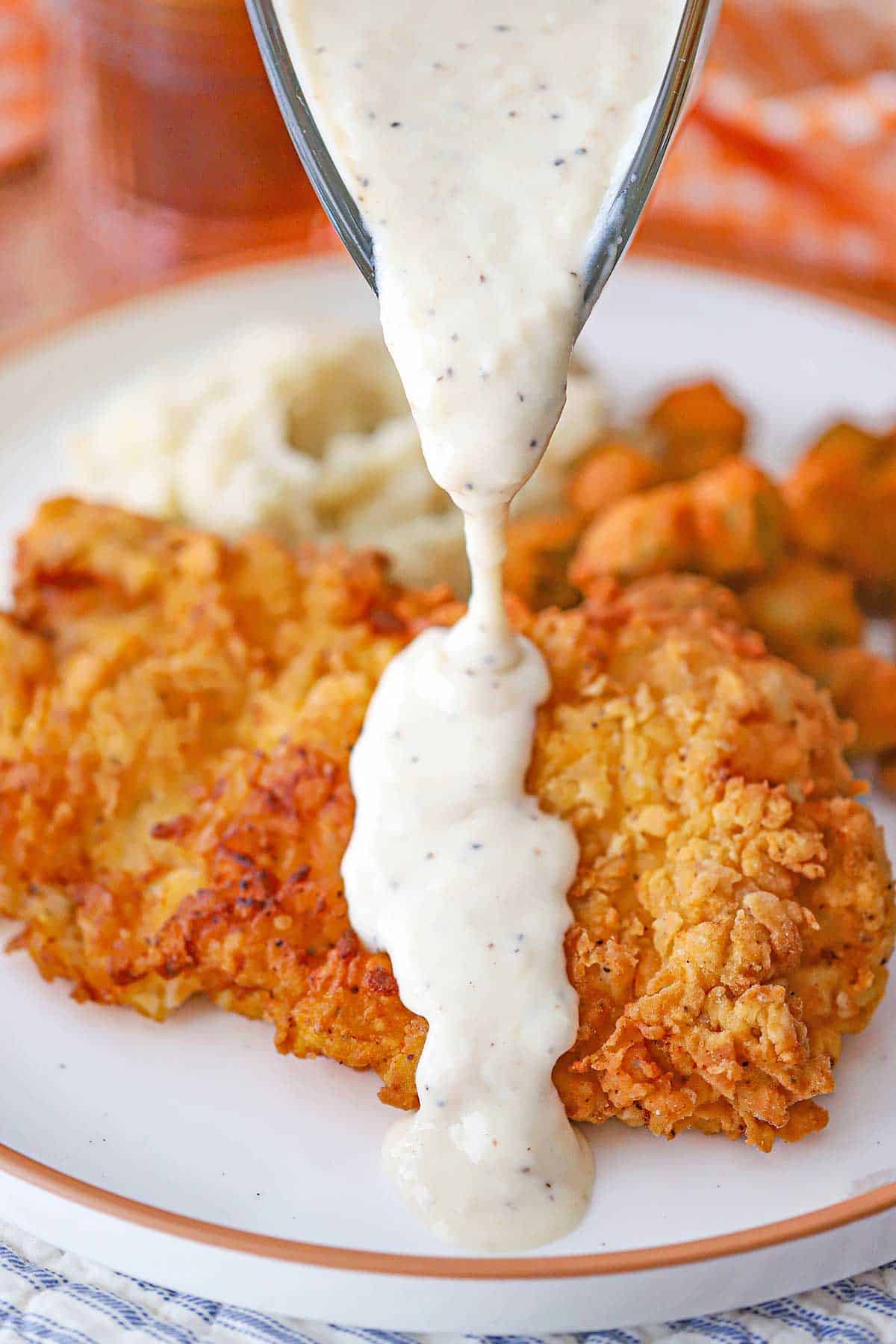 A person pouring Classic Southern Cream Gravy from a glass gravy boat over a cutlet of chicken fried chicken on a white dinner plate.