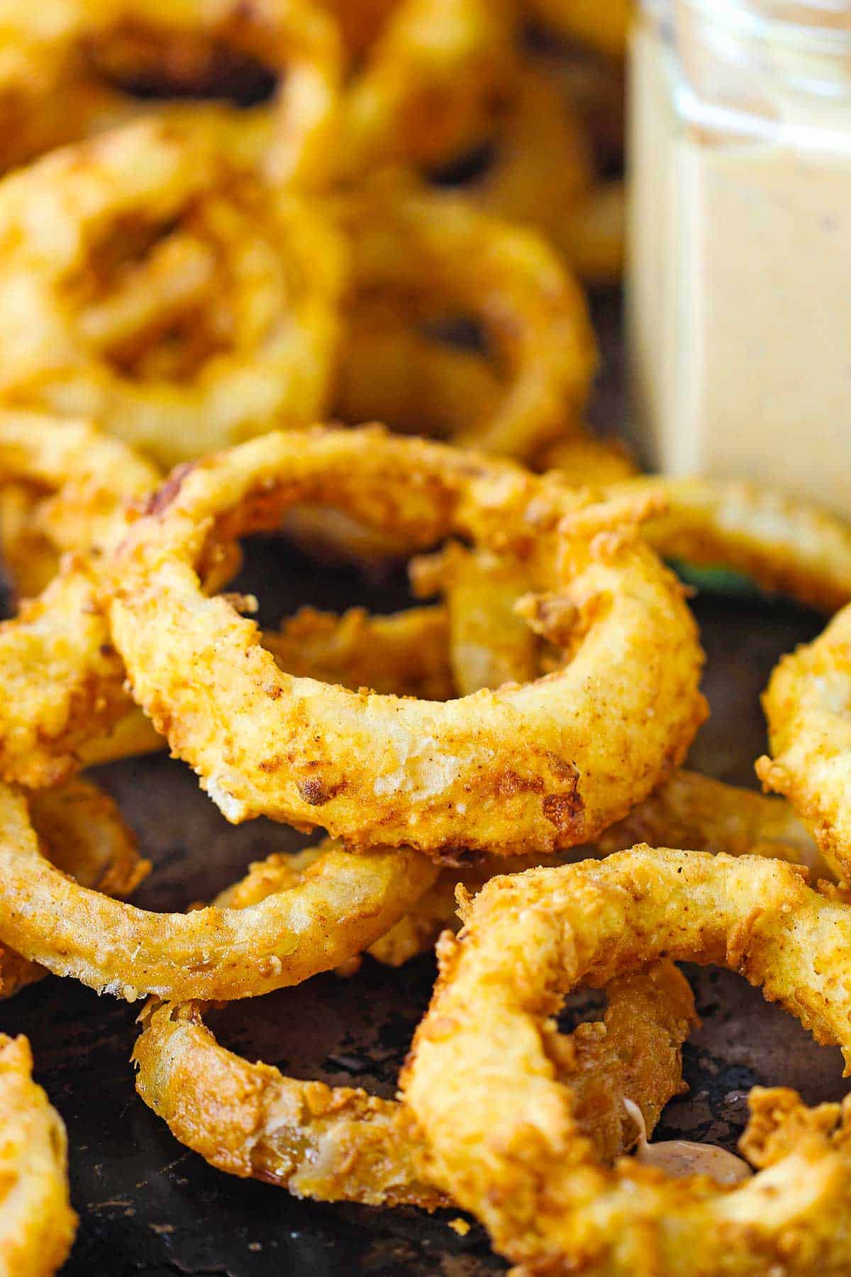 A close-up view of a pile of Steakhouse Onion Rings on a metal sheet pan next to a jar of Cajun Remoulade