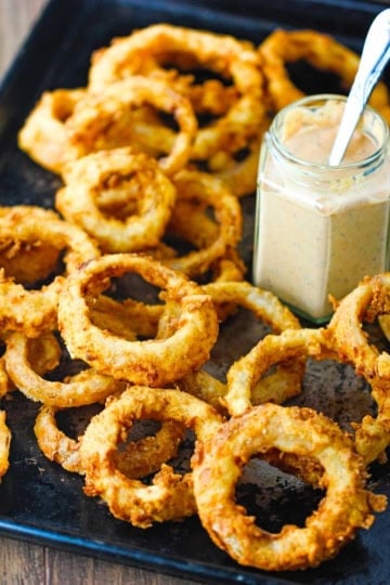 A pile of steakhouse onion rings on a baking pan with a jar or Cajun remoulade also on the pan.