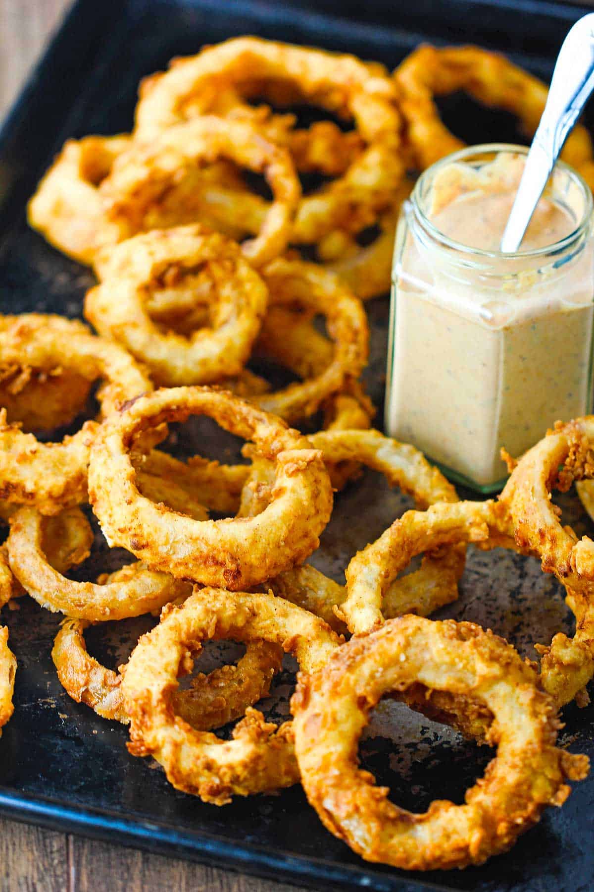 A pile of steakhouse onion rings on a baking pan with a jar or Cajun remoulade also on the pan.