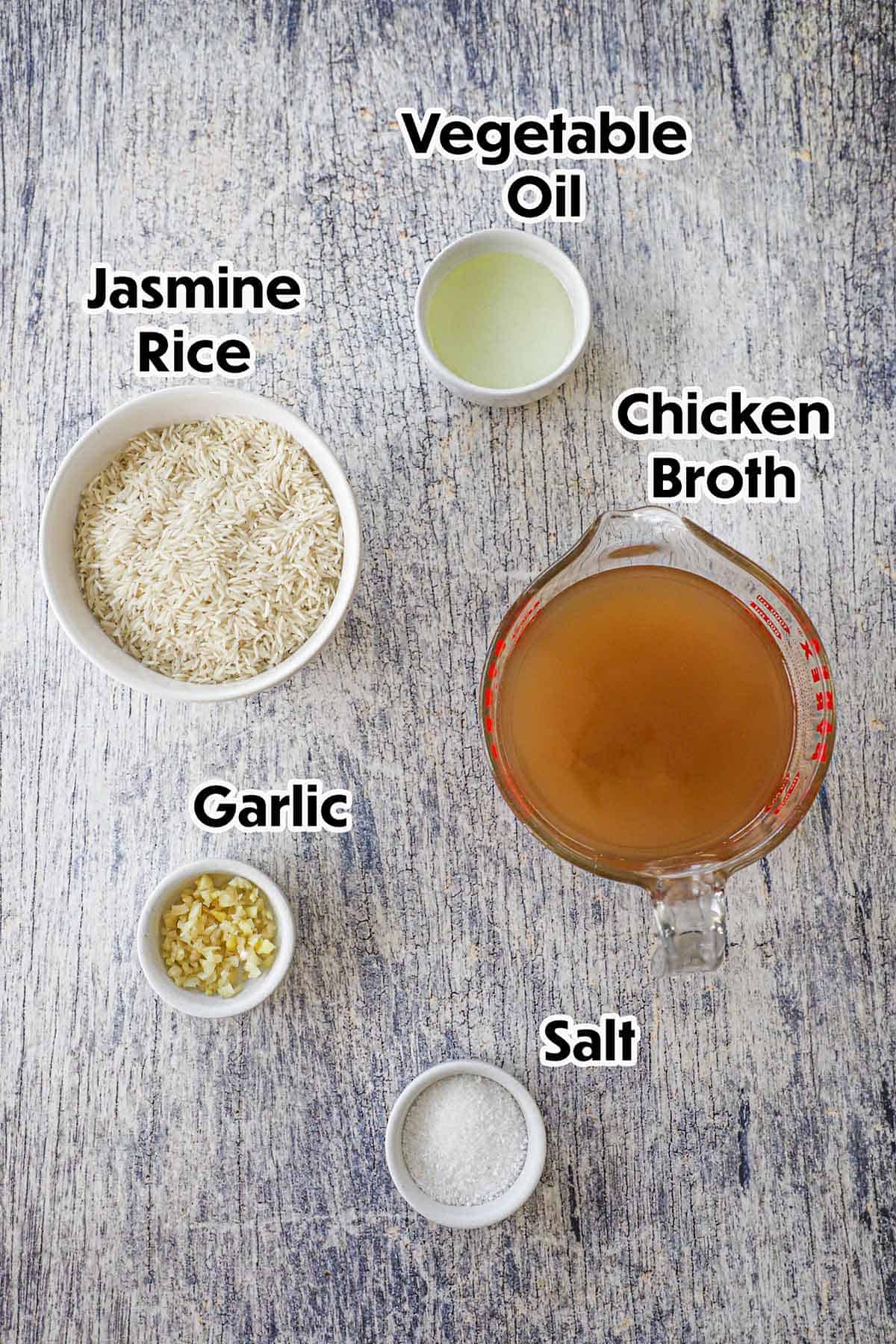 An arrangement of ingredients for Peruvian Garlic Rice on a grey wooden background including Jasmine rice, chicken broth, oil, garlic, and salt.