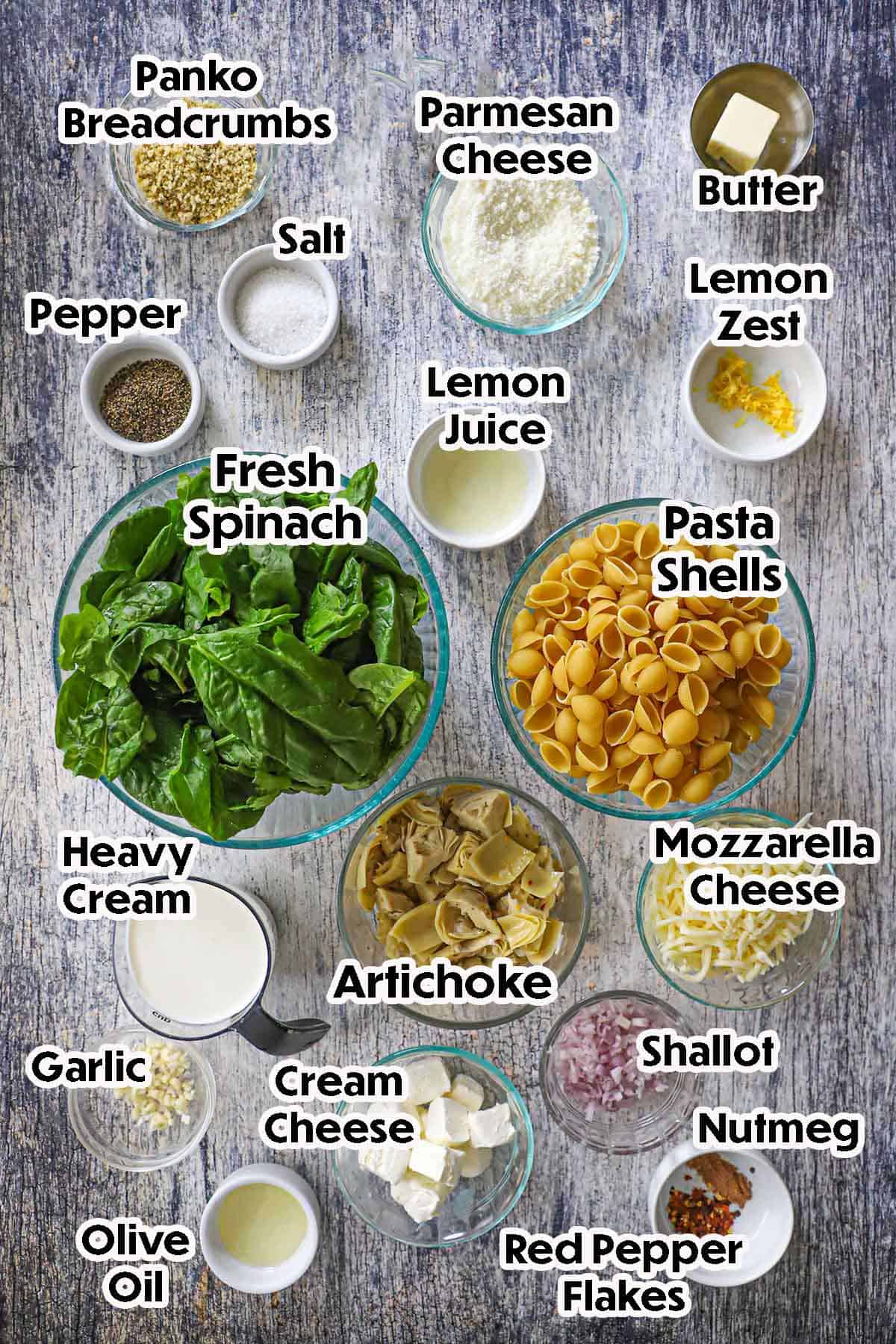 An arrangement of ingredients for Spinach Artichoke Dip Pasta on a grey wooden background including pasta shells, fresh spinach, artichokes, Mozzarella cheese, Parmesan cheese, lemon zest, heavy cream, cream cheese, shallot, garlic, and seasonings.