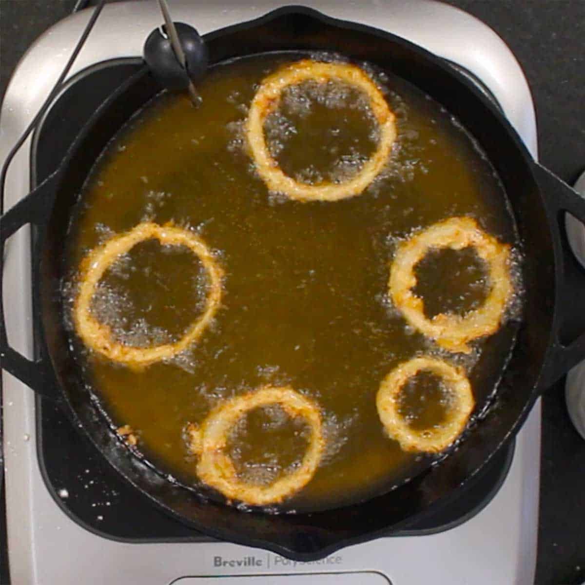 An overhead view of steakhouse onion rings being fried in hot oil in a large cast-iron skillet.
