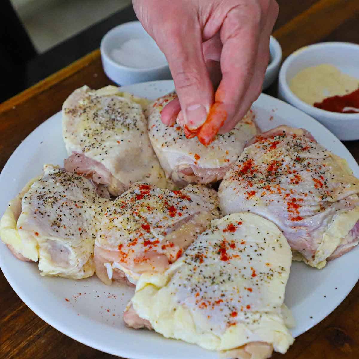 A person using his fingers to sprinkle smoked paprika over the top of raw chicken thighs that have been seasoned with salt and pepper.