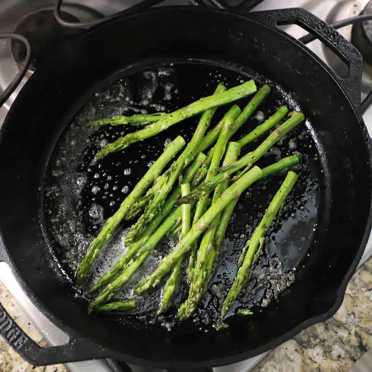 A bunch of seasoned asparagus stalks being sautéed in butter in a large black cast-iron skillet on a gas stove.