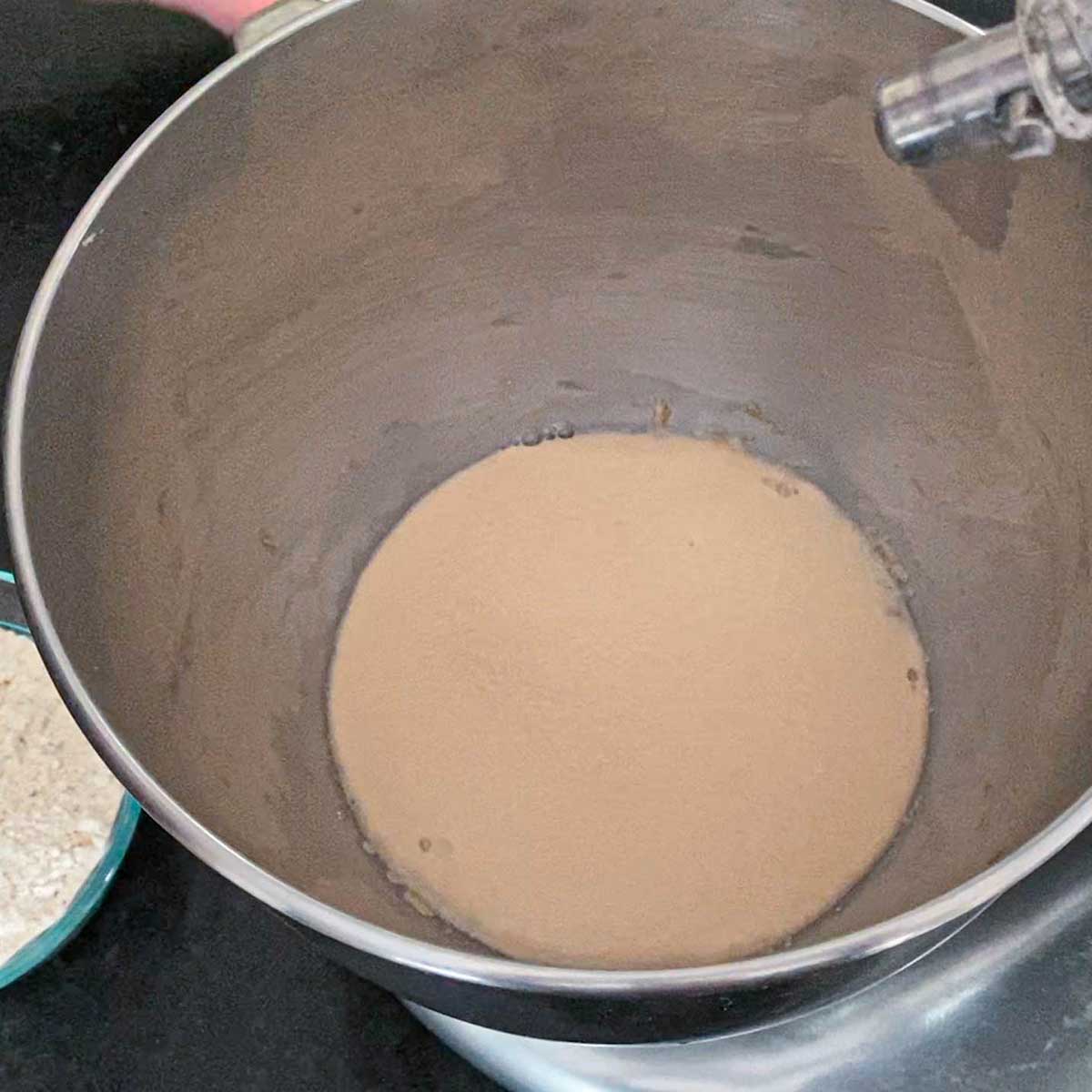 An overhead view of a metal mixing bowl filled with a mixture of active yeast that is blooming in warm water mixed with sugar.