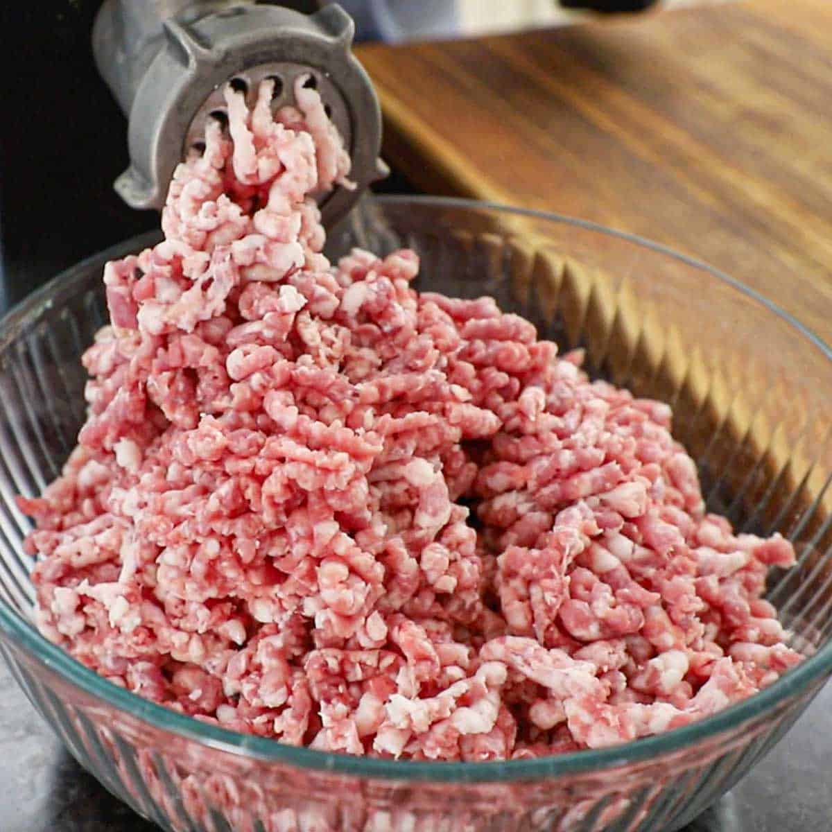 Pork butt being processed and ground through an electric grinder into a large glass bowl.